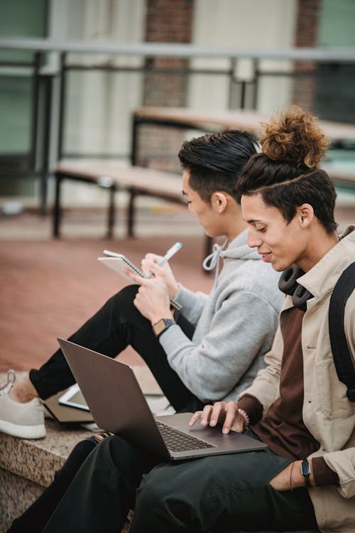 Multiethnic male students sitting with computer and notebook in street