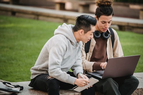 Young multiethnic male students in casual outfit sitting on stone border near green grass while using with computer and notebook in city street in daylight