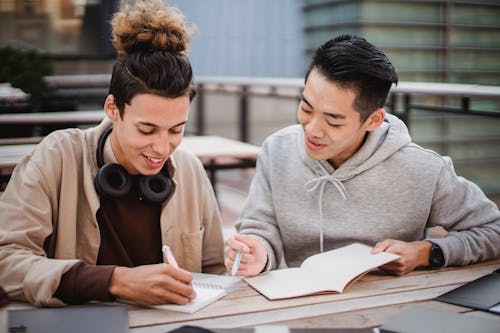 Free Multiethnic men students in casual clothes making notes in notepad while sitting at table in street in daytime Stock Photo
