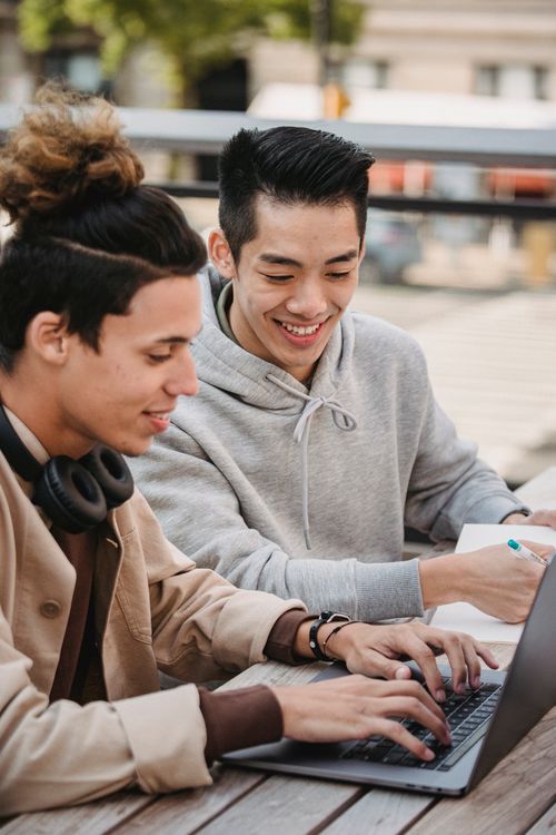 Free Smiling young multiracial male friends in casual outfit taking notes in notebook while using computer and sitting at table in street in daylight Stock Photo