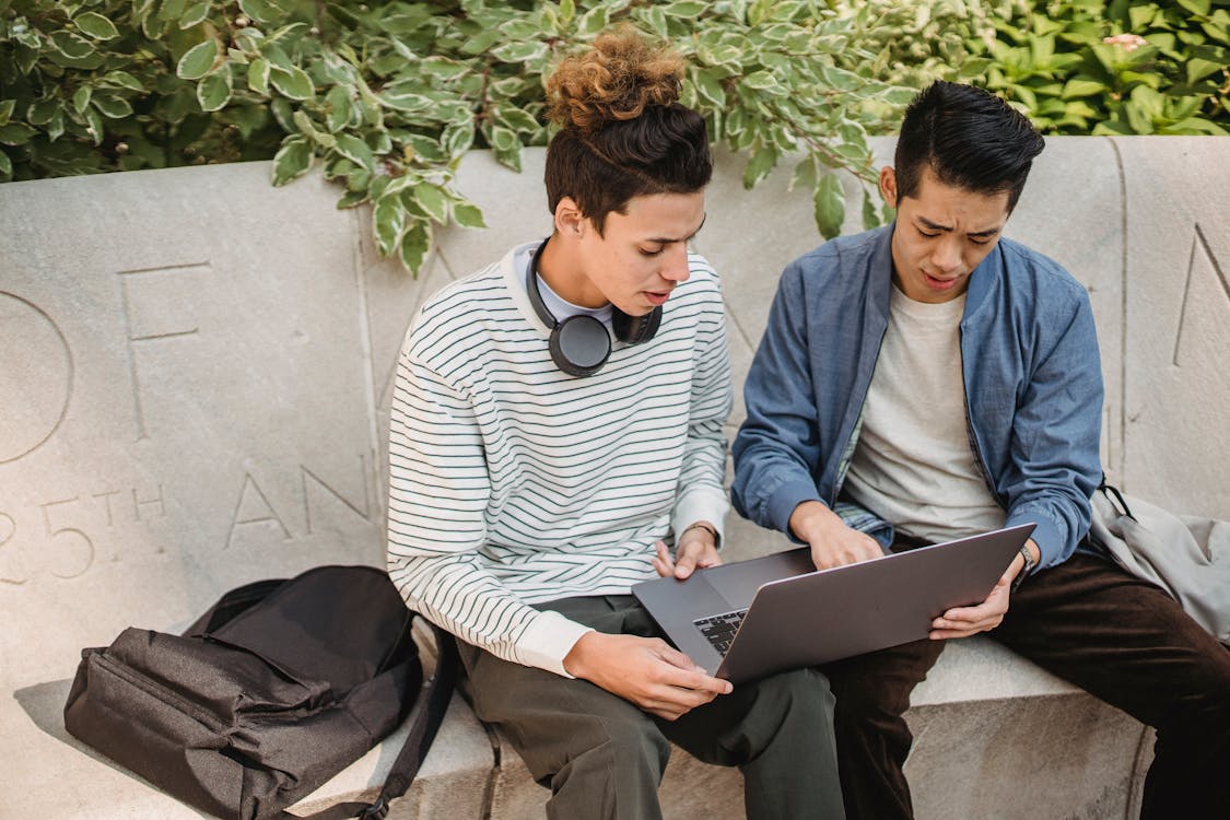 Serious young multiethnic men students in casual clothes studying on laptop while sitting on stone bench in street in daytime with backpack near green plants