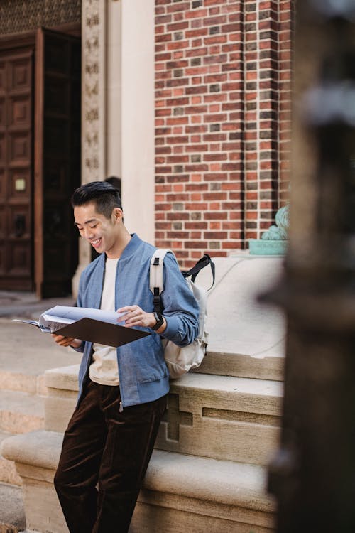 Free Smiling young Asian male in casual clothes and backpack standing in street near building while reading folder with papers in daytime Stock Photo