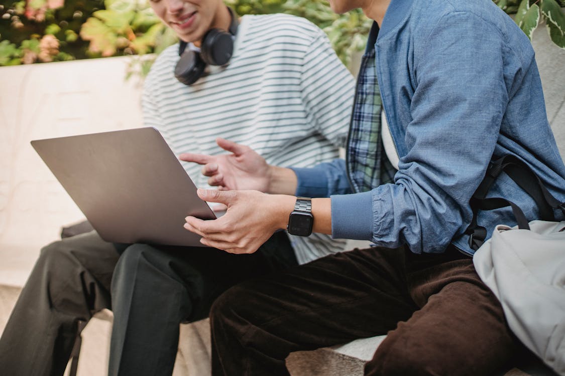 Crop faceless men students in summer outfit surfing on computer while sitting on stone bench in daytime in street