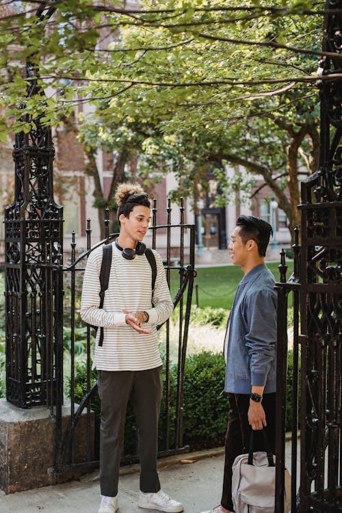 Young multiethnic men in casual outfit with backpacks standing in city street near fence with building and having conversation in daylight