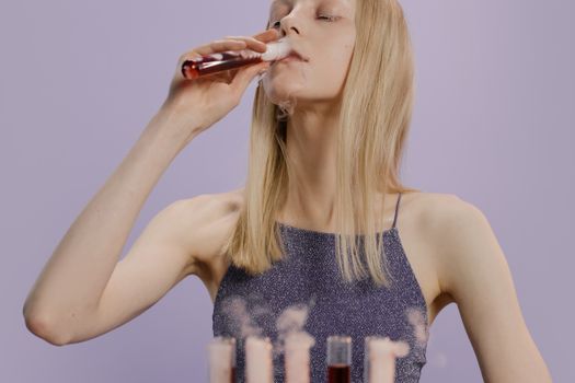 一位身穿灰色连衣裙的白人妇女从紫色背景中的彩色液体试管里喝水