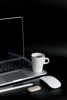 A sleek minimalist setup featuring a laptop, coffee cup, and gadgets on a black background.
