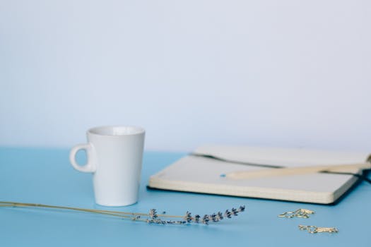 A serene minimalist workspace features a ceramic cup, notebook, lavender flower, and office supplies.