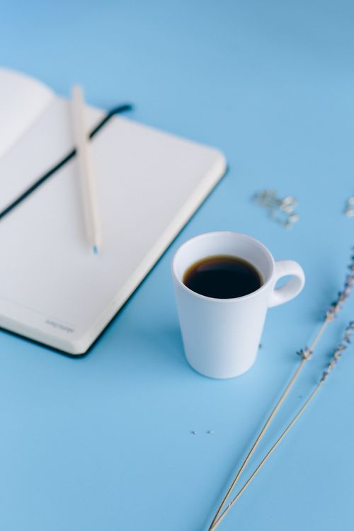 Free White ceramic mug of coffee with a notebook and pencil on blue background. Perfect for productivity themes. Stock Photo