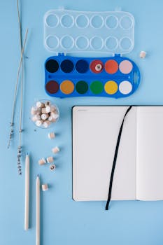 Artistic flat lay featuring watercolor paints, notebook, and pencils on blue background.