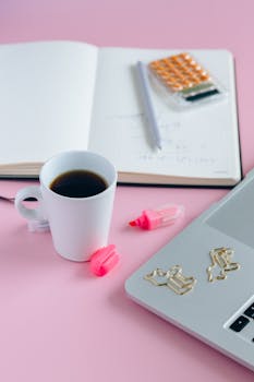 Organized workspace setting with coffee, stationery, and laptop on a pink surface.