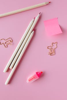 Flat lay of colorful stationery including pencils and clips on a pink surface, perfect for creative projects.