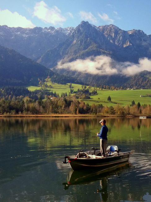 A fisherman enjoys a tranquil morning fishing on a lake surrounded by towering mountains, under a beautiful sky.