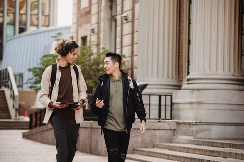 Free Young multiethnic men students in casual clothes strolling in city near building on pavement with folder and having conversation in daytime Stock Photo