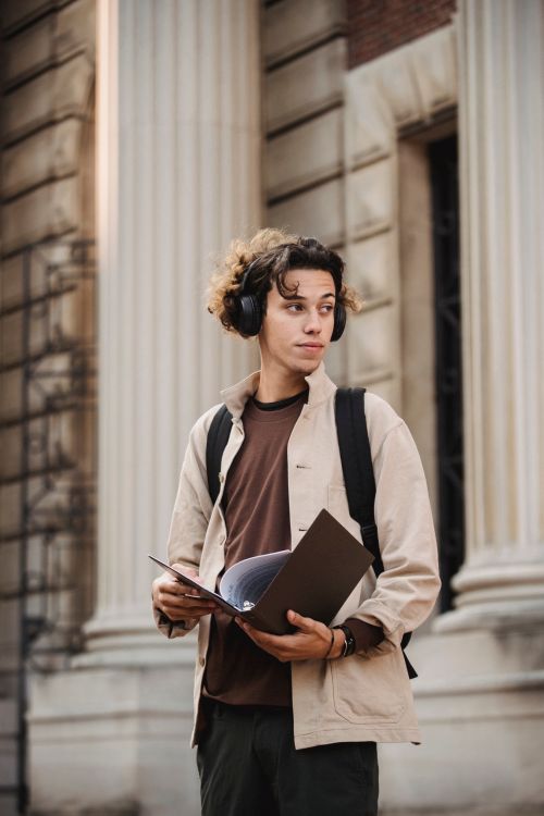 Free Young ethnic man in casual outfit and headphones with backpack standing in street near building with folder and papers in day Stock Photo