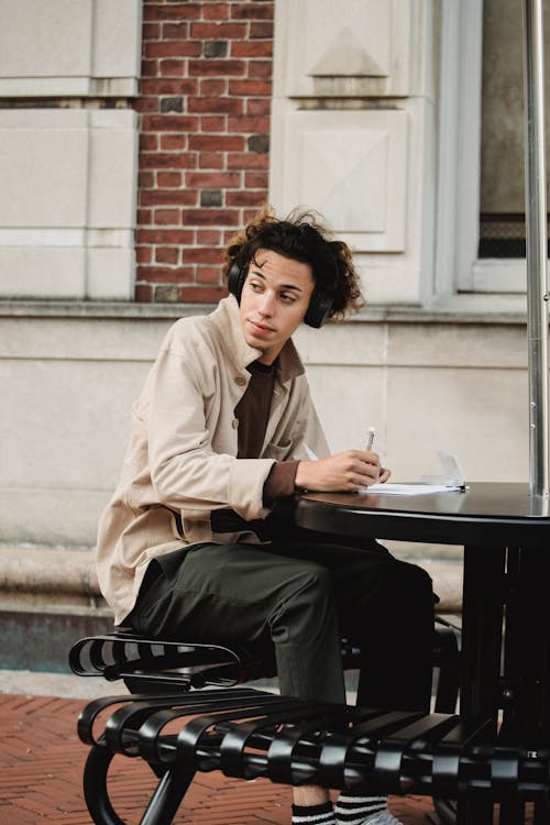 Serious young ethnic male in casual clothes and headphones sitting on bench at table outside and writing information in papers with pen in day and looking away
