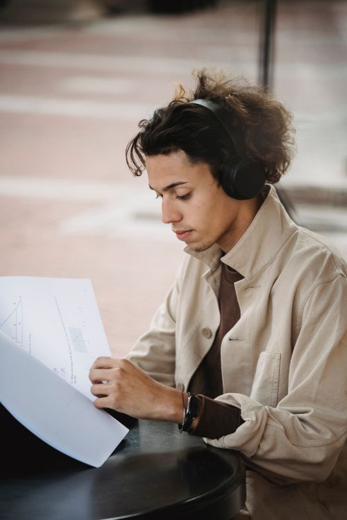 Free Young ethnic male in casual clothes and headphones sitting in street at table and reading papers in daytime Stock Photo