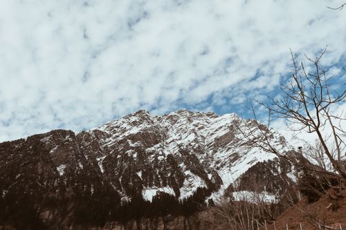 Free Picturesque view of snowy mountain range near hill with leafless trees under blue cloudy sky Stock Photo