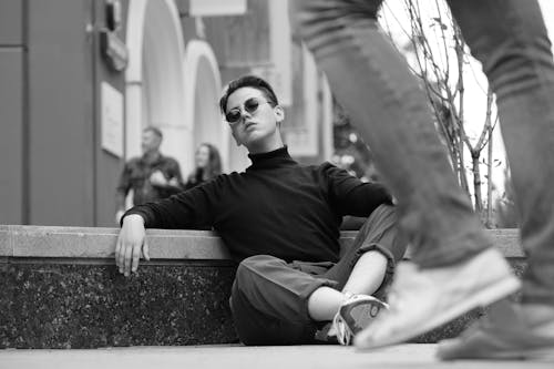 Free Black and white young arrogant male in stylish clothes and sunglasses sitting confidently on road curb on street and looking at camera with challenging glance Stock Photo