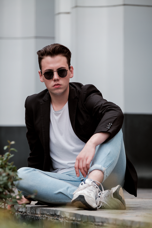 Free Young confident male in trendy wear and black sunglasses sitting with legs crossed on paved sidewalk outside modern building and looking at camera with attitude Stock Photo
