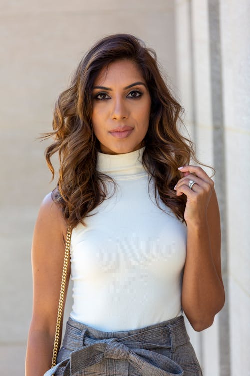 Young female with wavy hair in classy outfit looking at camera near gray wall of building