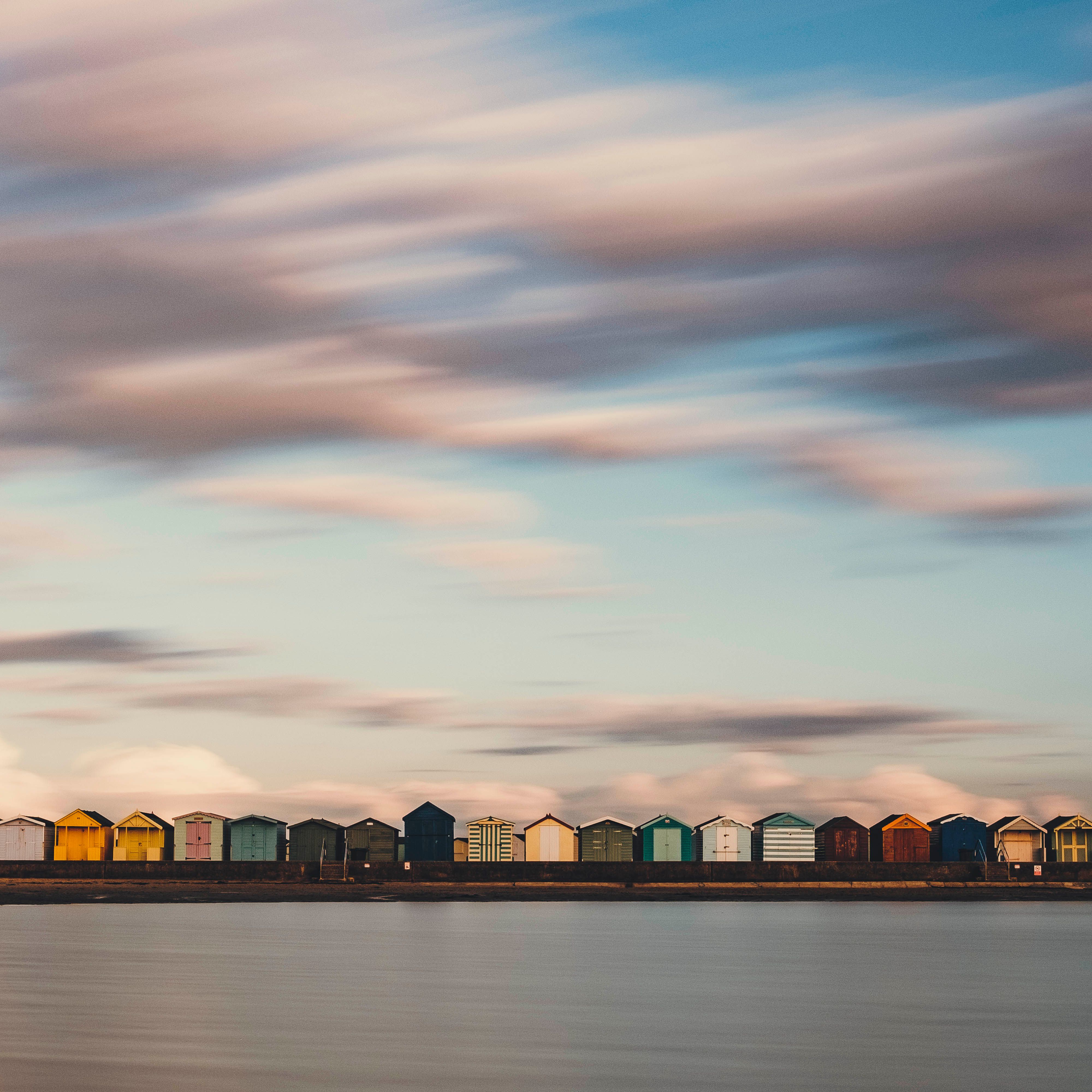 Fotomural Larga exposición de vibrantes cabañas en la playa al anochecer con mar suave y movimiento de nubes.