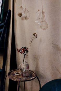 乡村场景中，一个玻璃花瓶里插着干花，上方悬挂着复古装饰的灯泡