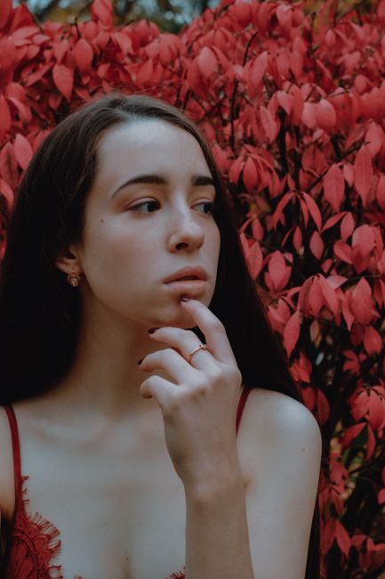 Young wistful female in trendy wear touching chin while looking away near red bush