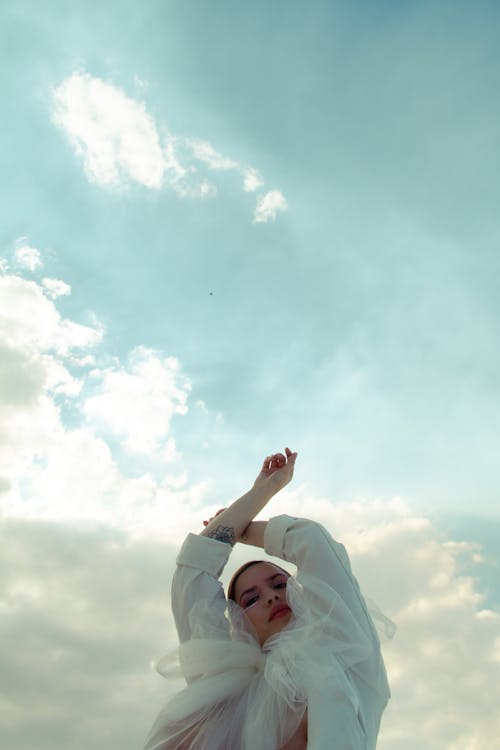 From below of graceful female in trendy outfit raising arms and looking at camera under cloudy sky