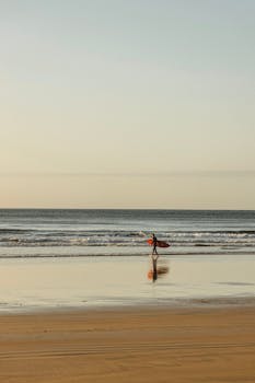 A lone surfer in a wetsuit walks along Bells Beach reflecting the dawn sky.