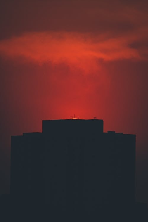 Silhouette of contemporary building located under red sky with floating cloud at sunset time in city in night in darkness