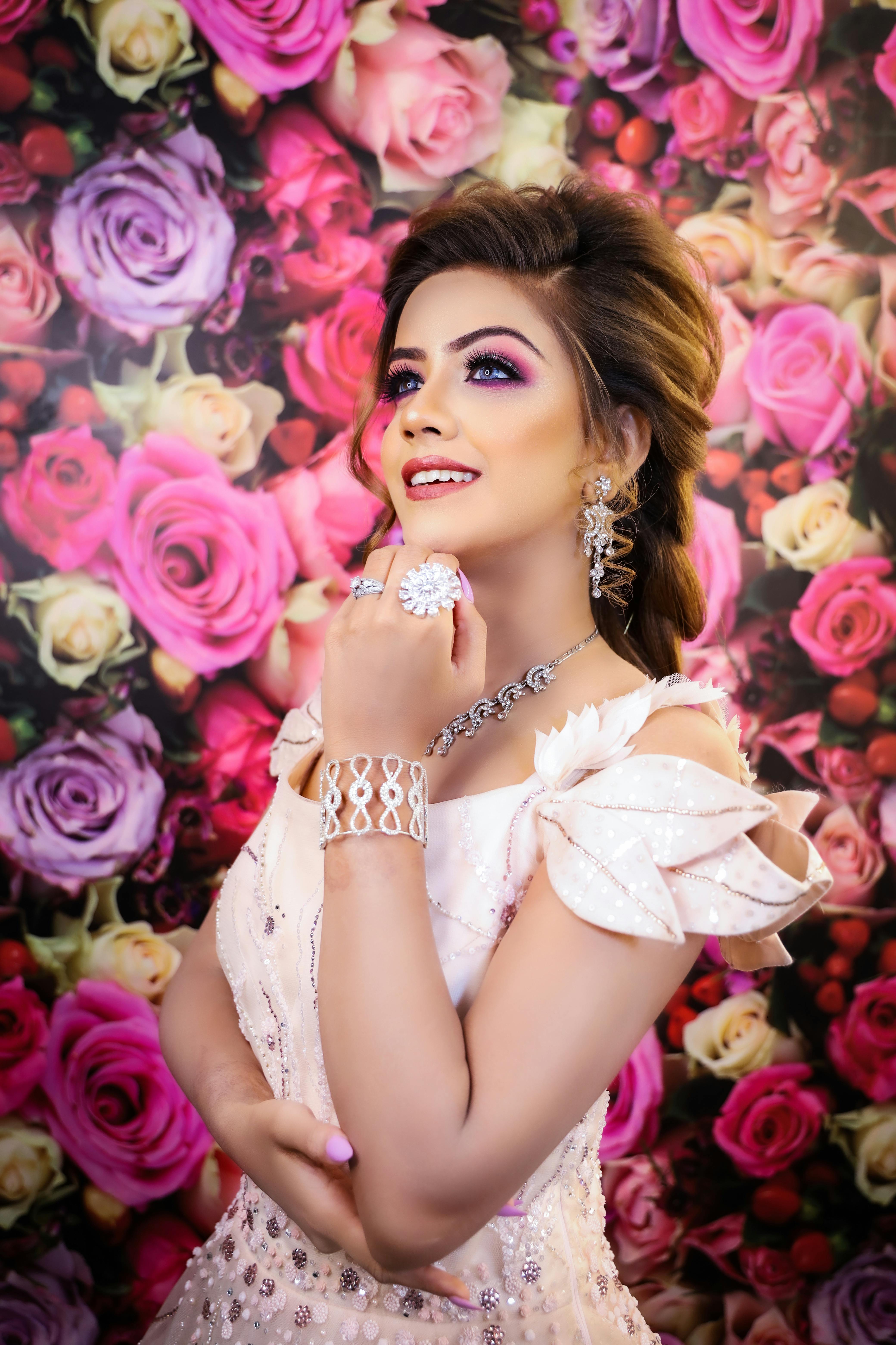 Dreamy young woman in pink dress with hairdo in makeup with earrings and necklace with rings and bracelet looking away happily near wallpapers covered with roses