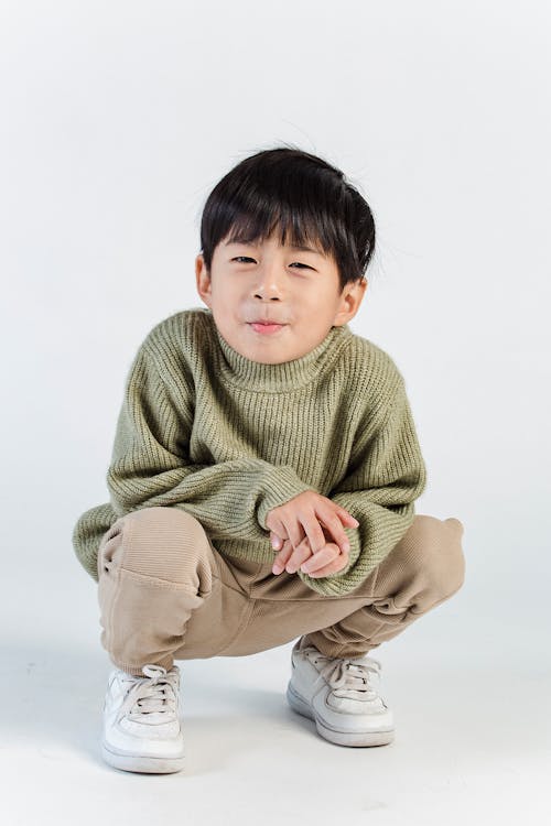 Full body of adorable Asian child wearing trendy casual wear sitting on hunkers against white background