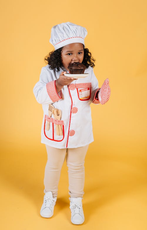 Full body of small African American child wearing chef uniform and oven gloves eating yummy muffin