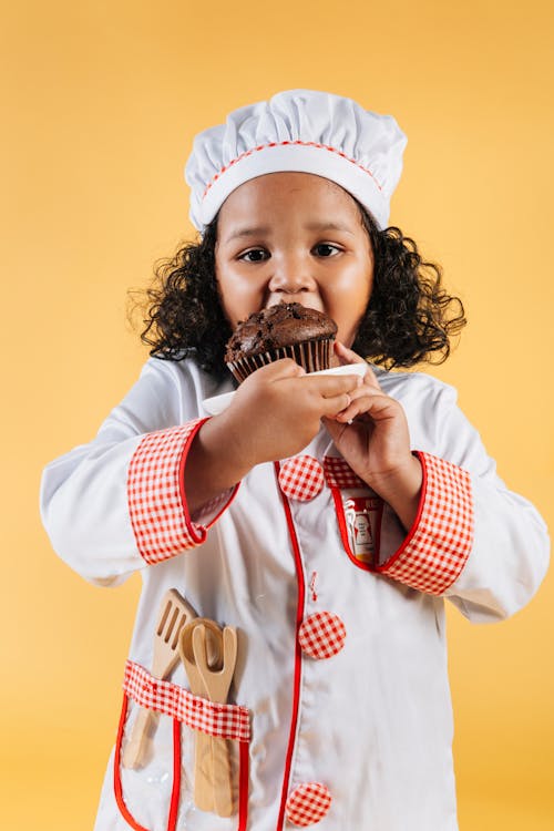 Cute African American child wearing chef wear eating taste sweet chocolate dessert in yellow studio