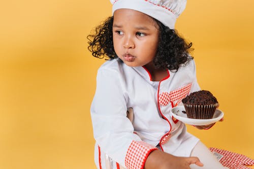 Adorable African American girl in chef uniform and hat eating muffin against yellow background