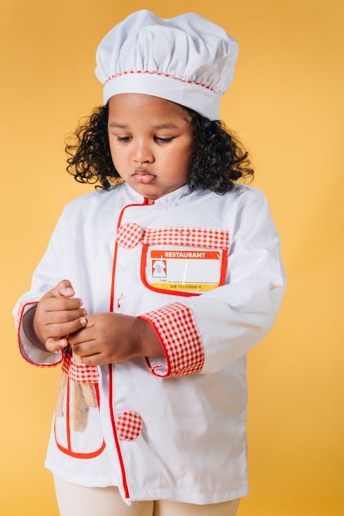 Curios African American child wearing chef uniform and hat standing with kitchen tools against yellow background