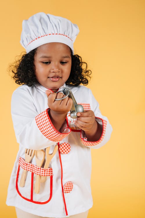 Adorable funny little African American girl looking at spoons attentively on yellow background of studio