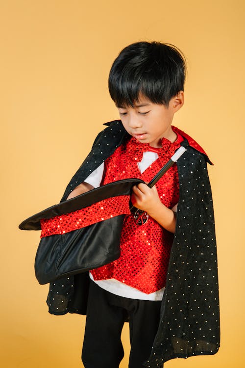 Cute funny ethnic boy in costume of magician with magic wand looking at hat with interest on yellow background