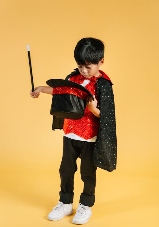 Free Full body of adorable magician Asian boy wearing cape standing on yellow background with wand and looking into black hat in studio Stock Photo