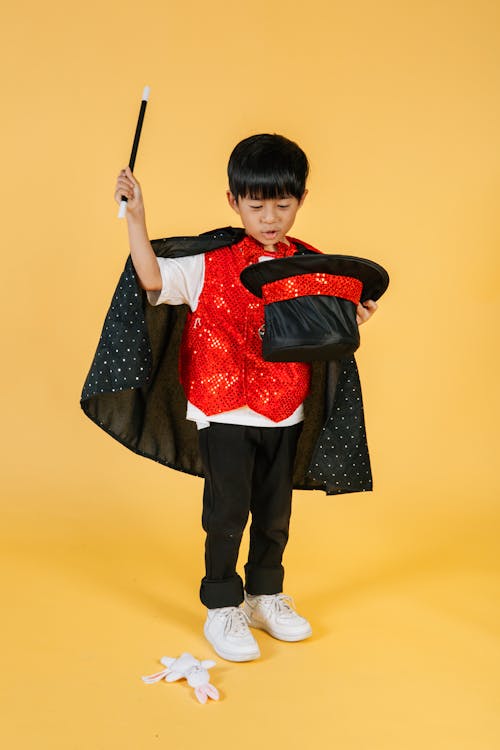 Full body of little ethnic boy in magician costume with cape doing magic trick while using cylinder hat and magic wand while standing on orange background