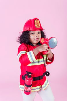 身穿消防员服装、头戴带灭火器头盔的民族孩子，站在粉红色背景前，手持假扩音器