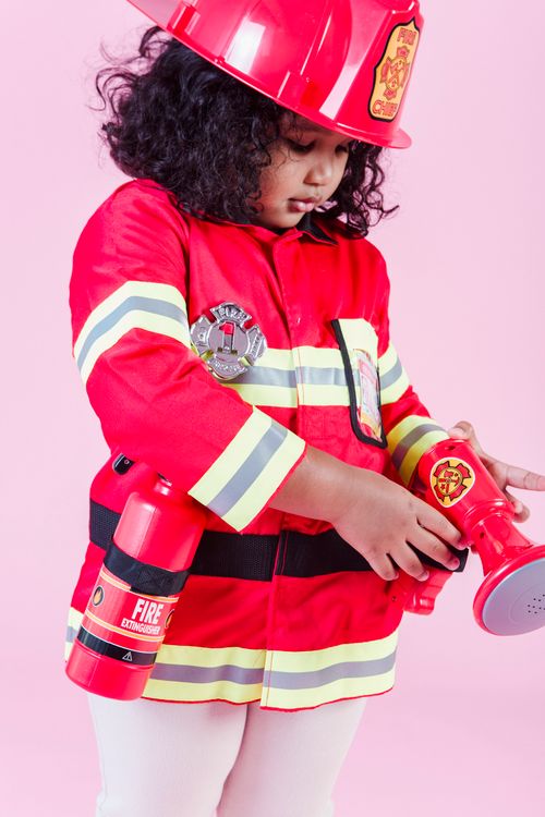 Free Ethnic kid in firefighter costume with fire extinguisher and hardhat using megaphone toy while standing on pink background Stock Photo