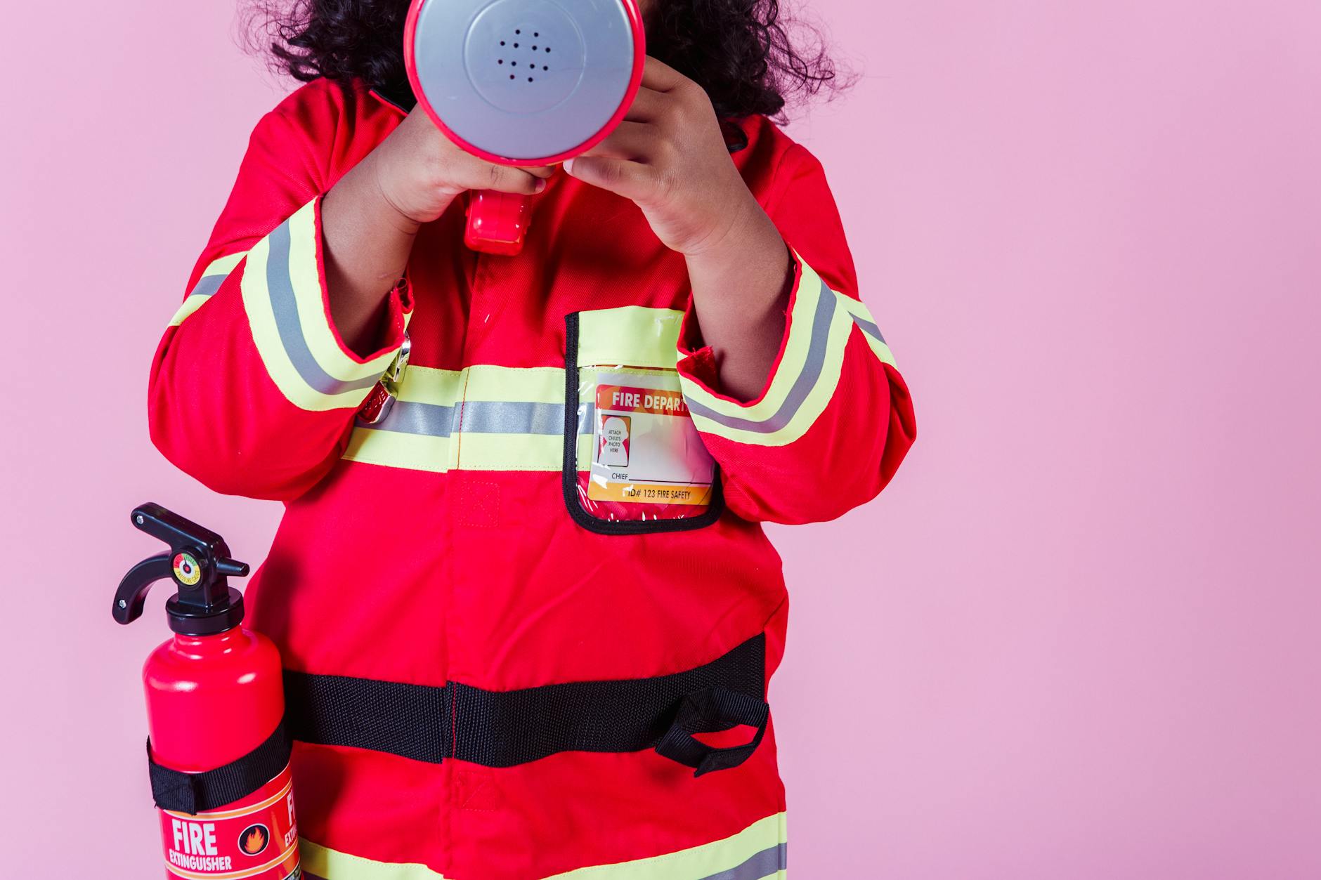 aumentar la visibilidad Crop unrecognizable little ethnic kid wearing fireman costume and fire extinguisher while using loudspeaker toy and standing on pink background