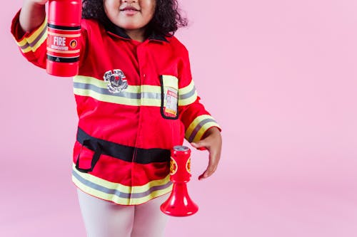 Crop anonymous little ethnic kid in fireman costume with fake fire extinguisher and megaphone standing on pink background