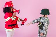 Side view of multiracial children in military uniform and fireman costume with megaphone and fire extinguisher standing together on pink background in helmets