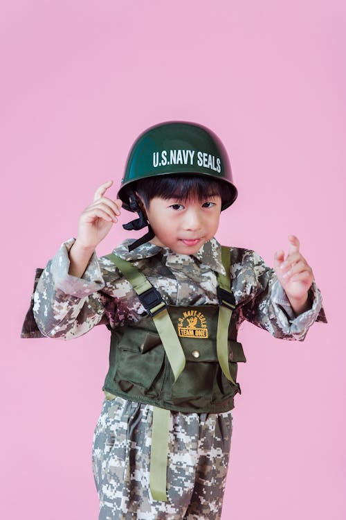 Ethnic boy in military costume with hardhat standing on pink background and looking at camera with raised hands