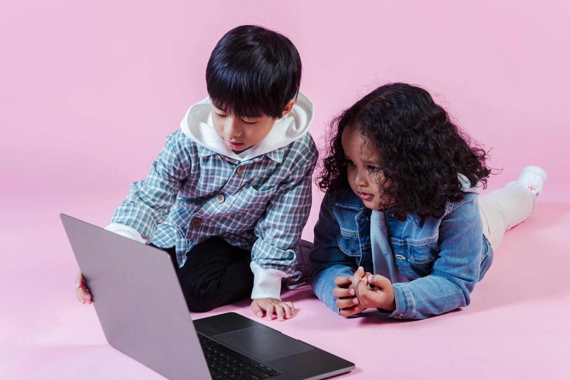 Little multiracial children in casual outfit spending time on floor while using netbook on pink background