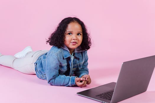 A young girl with curly hair uses a laptop while lying on a pink background, depicting modern digital interaction.