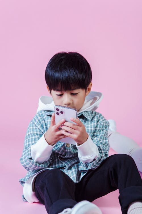Free Little Asian kid in casual outfit sitting on floor and using smartphone on pink background Stock Photo