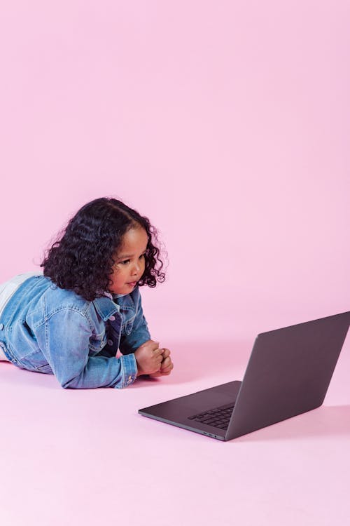 Free Little ethnic girl in casual denim jacket lying on floor with laptop on pink background Stock Photo